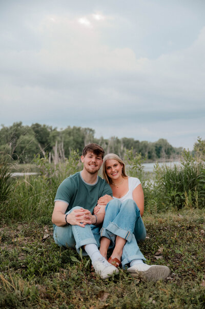 Couple poses for the camera on the grass near a lake smiling at the camera.
