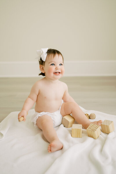 Baby boy with blue eyes in white diaper cover sits up and chews on wooden toy during baby photography session in Raleigh NC studio