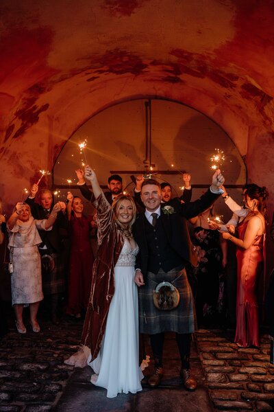 A couple are photographed surrounded by their wedding guests. The stand in a pink archway and all hold sparklers.