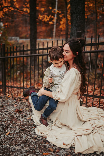 Mother holds son in her lap outside in front of a fence surrounded by colorful leaves in the fall