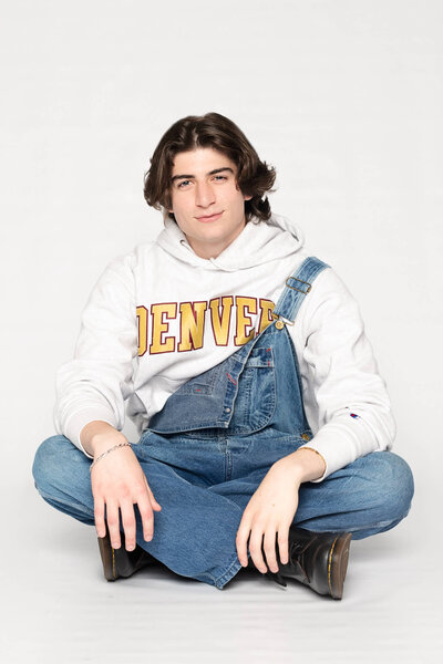 A dark haired young man wearing jeans and a light green sweatshirt jumping in the air in a white studio