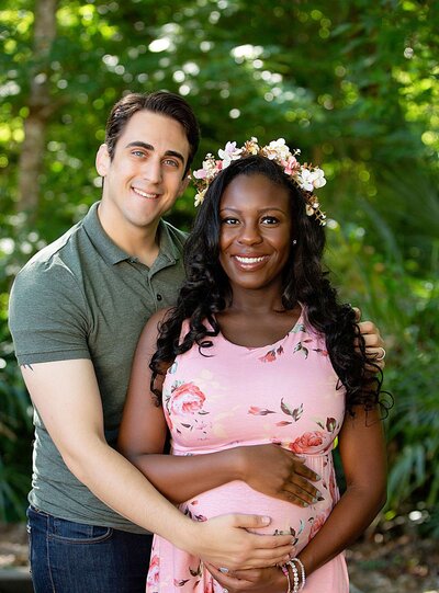 A pregnant couple smiles together for a photo, surrounded by lush greenery in a serene woodland setting.