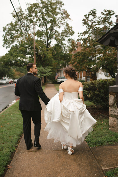 Bride and groom walk hand-in-hand down a neighborhood sidewalk, the bride lifting her dress as they smile at each other on a cloudy wedding day.