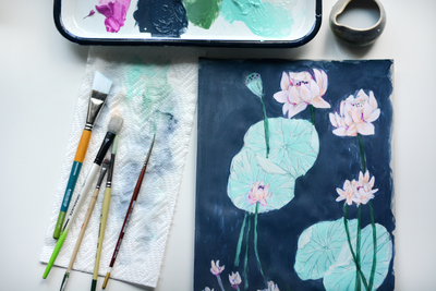 An overhead image of paint brushes and a palette with a botanical painting of lily pads, from the desk of Skye McNeill. Read the blog.