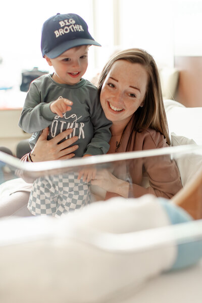 Mother and toddler looking into a hospital cot at newborn baby brother, with the toddler pointing excitedly at the baby