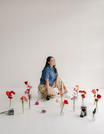 A woman in a denim shirt and floral skirt kneels on a white floor, surrounded by vases of red and pink flowers. She holds a camera, smiling peacefully.