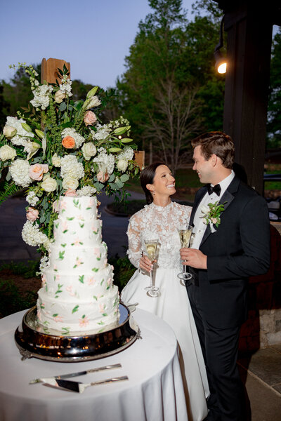 white cake with flower arrangement behind it and couple toasting next to it