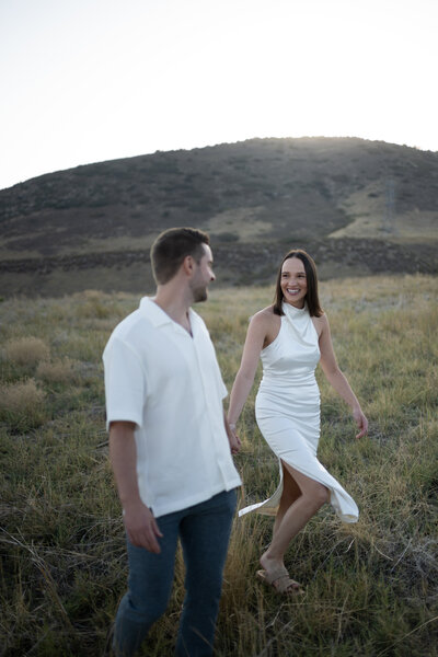 A couple walks hand in hand through a golden grassy field at sunset, surrounded by rolling hills. The woman, wearing a sleek white halter dress with a thigh slit, smiles warmly at her partner. Her dark hair is neatly styled, and she moves gracefully through the tall grass in strappy sandals. The man, dressed in a casual white short-sleeved button-up and dark jeans, gazes back at her with admiration. The soft, fading sunlight casts a dreamy glow over the landscape, capturing a moment of effortless romance and joy.