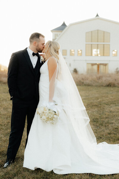 Couple kissing in front of white barn venue.