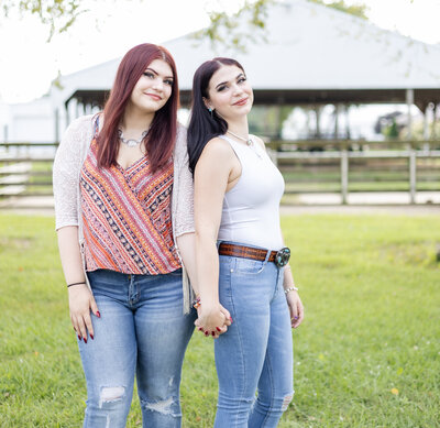 Sisters standing next to each other in the grass, looking at the camera with a soft smile, holding hands.  One sister has long red hair, wearing blue jeans, a light tan smock over a patterned shirt.  Other sister has long black hair, wearing blue jeans, brown belt, and a white sleeveless shirt.