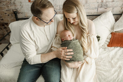 A serene family portrait featuring a mother and father seated together, lovingly holding their newborn baby. The baby is peacefully swaddled in a cozy green knit blanket. The mother wears a soft, off-the-shoulder cream dress, and the father, in a casual cream shirt and glasses, gazes at the baby with affection. The neutral tones and textured backdrop add warmth and intimacy to the image.