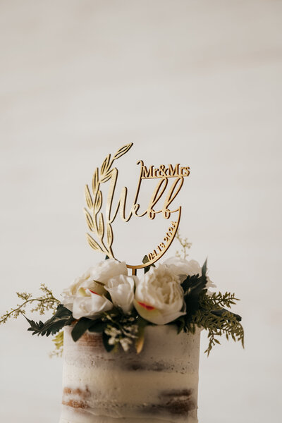 Naked cake with white flowers and a topper that says "Mr and Mrs Webb 04.13.2024"