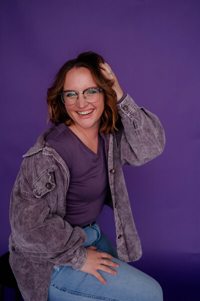 Woman poses for the camera with her hand in her hand wearing denim in front of a purple backdrop.