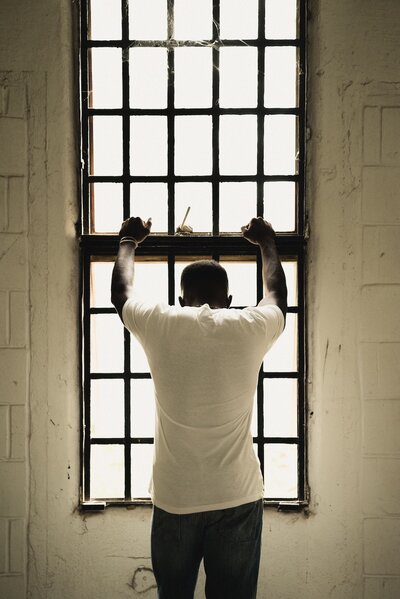 A man with tattoos on his arms wearing an orange jumpsuit inside a prison cell