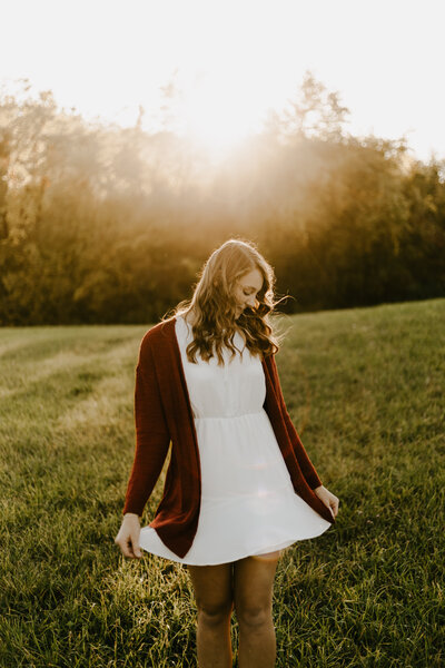 A senior standing in a grassy green field at sunset wearing a beautiful white dress. 