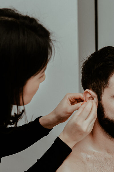 Woman with dark hair wearing a black shirt administers ear acupuncture on male patient with short, dark hair and beard,