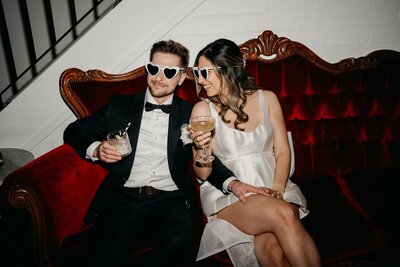 Bride and groom in heart sunglasses sharing drinks on a red velvet couch at Gracelands, a fun candid wedding photo on the Central Coast.