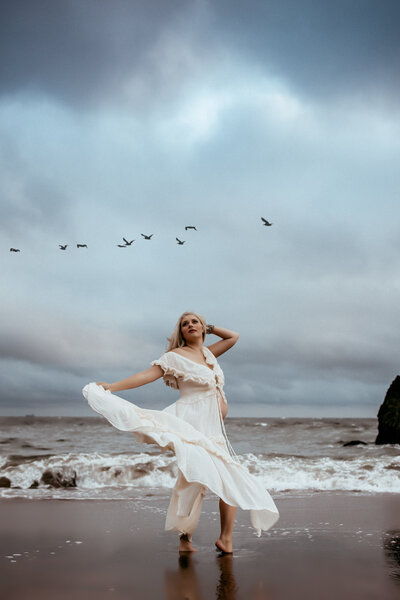 A blonde woman in a flowy white dress twirls barefoot on the beach while the seaguls fly overhead. 