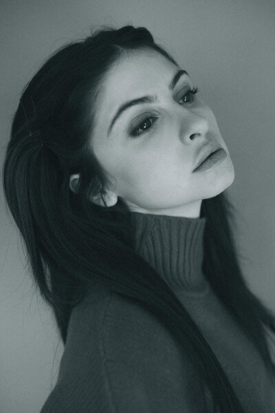Woman in a bright blue sweater and jeans poses in profile against a clean white wall, hands resting on her hips.