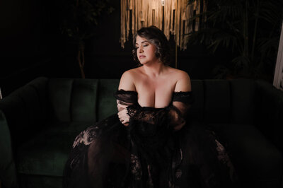 A woman in a black lace coverup sits on a green couch looking down her exposed shoulder