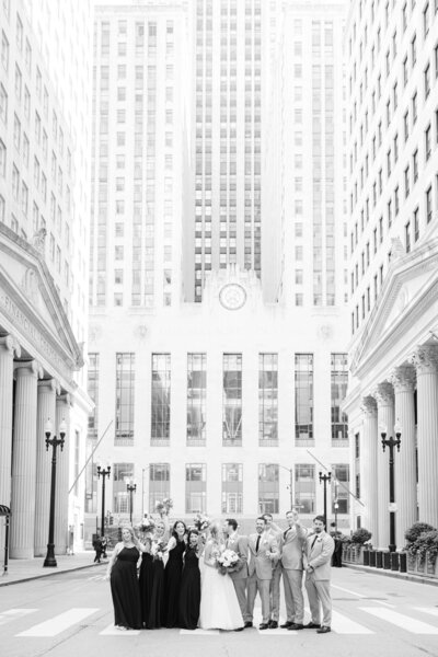 Black and white photo of a wedding party celebrating in the middle of LaSalle Street in front of the iconic Chicago Board of Trade building. The bride and groom stand at the center surrounded by bridesmaids in dark dresses and groomsmen in light suits, all smiling and cheering beneath the towering skyscrapers and classical architecture.
