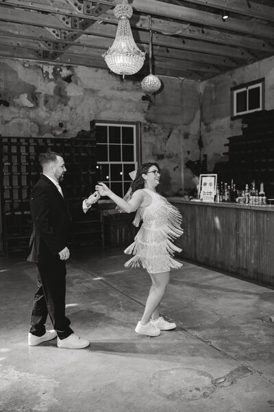 Black and white image of groom spinning bride while she dances in a short fringe dress
