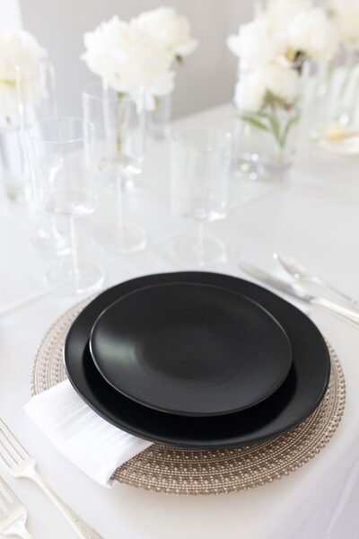 black plate and charger on top of a champagne colored large charger, white napkin, and white tableclothwith glass