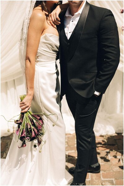 A closeup image of a bride standing with her groom holding her purple bouquet
