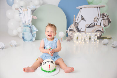 Smiling boy grabs cake with his fingers while sitting. There is a golf cart and bag in the background with balloon and arches.