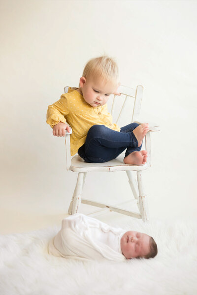 Toddler in a yellow shirt and jeans sitting in a chair looking at her newborn sister