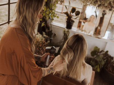 The best hair extensions professional in Frisco is holding up her flagship product. The professional has palm trees, leaves and leaves tattooed on her arms. Behind the hair extensions is a wall tapestry with flowers