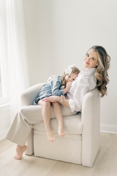 Newborn photographer, Meredith Maag, sits in a cream-colored chair with her young daughter laying across her lap, both barefoot and relaxed in a light-filled Kansas City studio