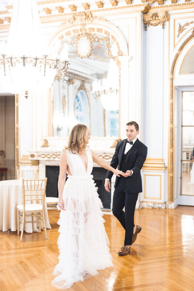 a bride reaches her hand back to hold the groom's hand as they walk through the music room of the elkins estate mansion near philadelphia. image by philadelphia wedding photographer emily wilkerson