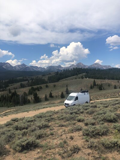 A white sprinter van drives along a road in Stanley Idaho with the Sawtooth Mountains in the background.