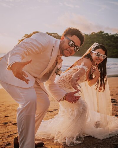 A bride and groom in stylish sunglasses strike a fun pose on the beach at sunset, celebrating their destination wedding in a lively and carefree way.