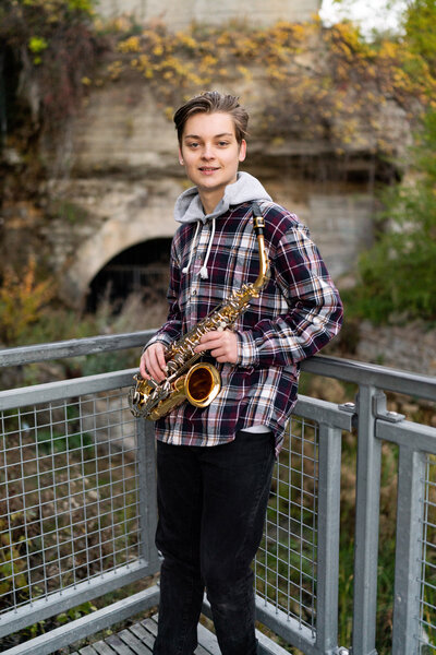 Senior boy in flannel holds saxophone while leaning into fence.