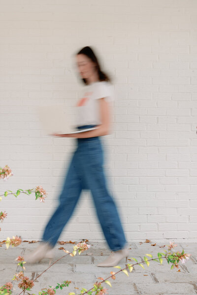 Blurred photo of a woman in a white top and blue jeans walking with her laptop in front of a white brick wall
