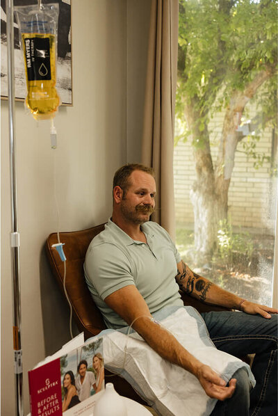 A man smiles as he is hooked up to an IV, receiving a peptide therapy treatment to help him achieve his wellness goals.