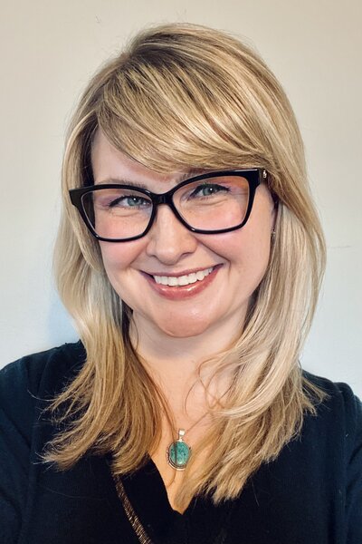 Vertical portrait of Kristin Tighe. She has blonde medium length hair, black glasses and is wearing a black shirt. 