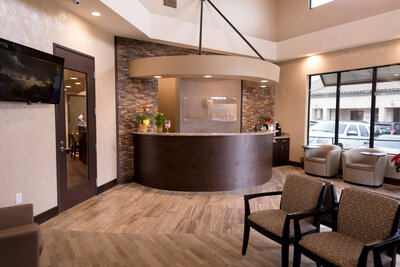 Transitional reception area in a dental office featuring a front desk, ledgestone wall, wood vinyl flooring, warm tones, and inviting atmosphere. The reception area is designed to create a welcoming and comfortable experience for patients. EnviroMed Design