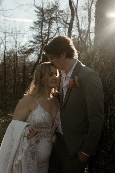 couple sharing an intimate moment during their golden hour forest elopement, with the groom gently kissing the bride’s forehead as she holds her embroidered lace jacket. The bride wears a deep V lace wedding gown, while sunlight filters through the trees, creating a romantic and natural backdrop perfect for an outdoor, adventure-inspired moment