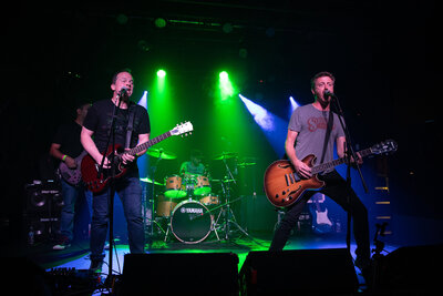 Under the vibrant green and purple stage lights, a band delivers an energetic performance. Two guitarists and vocalists captivate the front while a drummer keeps rhythm in the background. Capturing this live music magic requires a professional photography studio's skill for those perfect shots.
