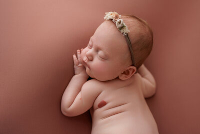 A newborn baby is peacefully sleeping on a soft, dusty rose-colored background. The baby is lying on their side with their tiny hands gently placed near their face. They are wearing a delicate headband adorned with small, fabric flowers in pastel shades of pink, cream, and green. The baby's expression is calm and content, with their eyes closed and their cheeks slightly rosy. A small birthmark is visible on the baby's arm, adding a unique touch to this serene and tender portrait.