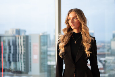 Woman wearing black suit coat standing in front of window in high rise apartment with the city in the boackground.