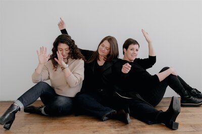 Portrait of Hudson Valley wedding planning team, Canvas Weddings, sitting  together and sharing a goofy moment in front of white wall