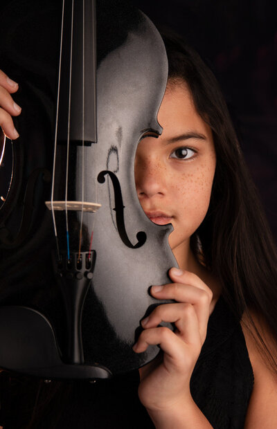 tween-girl-in-studio-arlington-tx-hol,ding-black-violin-over-one-eye-wearing-black-against-black-background