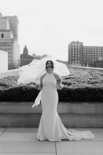 Black and white image of bride with veil blowing over her face while looking happy