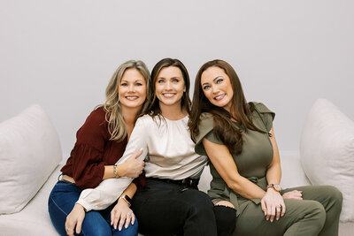 Three women are sitting on a white couch, smiling. One is wearing a burgundy top, another a white blouse, and the third a green outfit. They have long hair, styled differently, and are positioned closely together against a plain background.