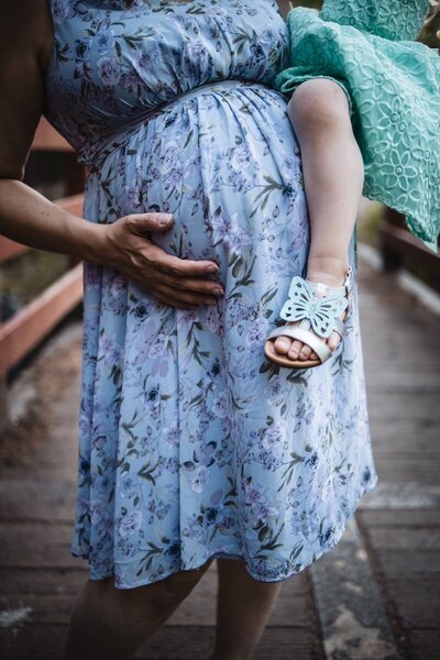 Pregnant mom in a blue floral dress holding her belly with one hand and her daughter in the other