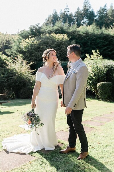 Bride and groom smiling at each other in a sunlit garden setting, with the bride in an off-the-shoulder gown and holding a wildflower bouquet during their countryside wedding